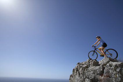 Female mountain biker sitting on bicycle at edge of rock in sunlight, looking at horizon over sea